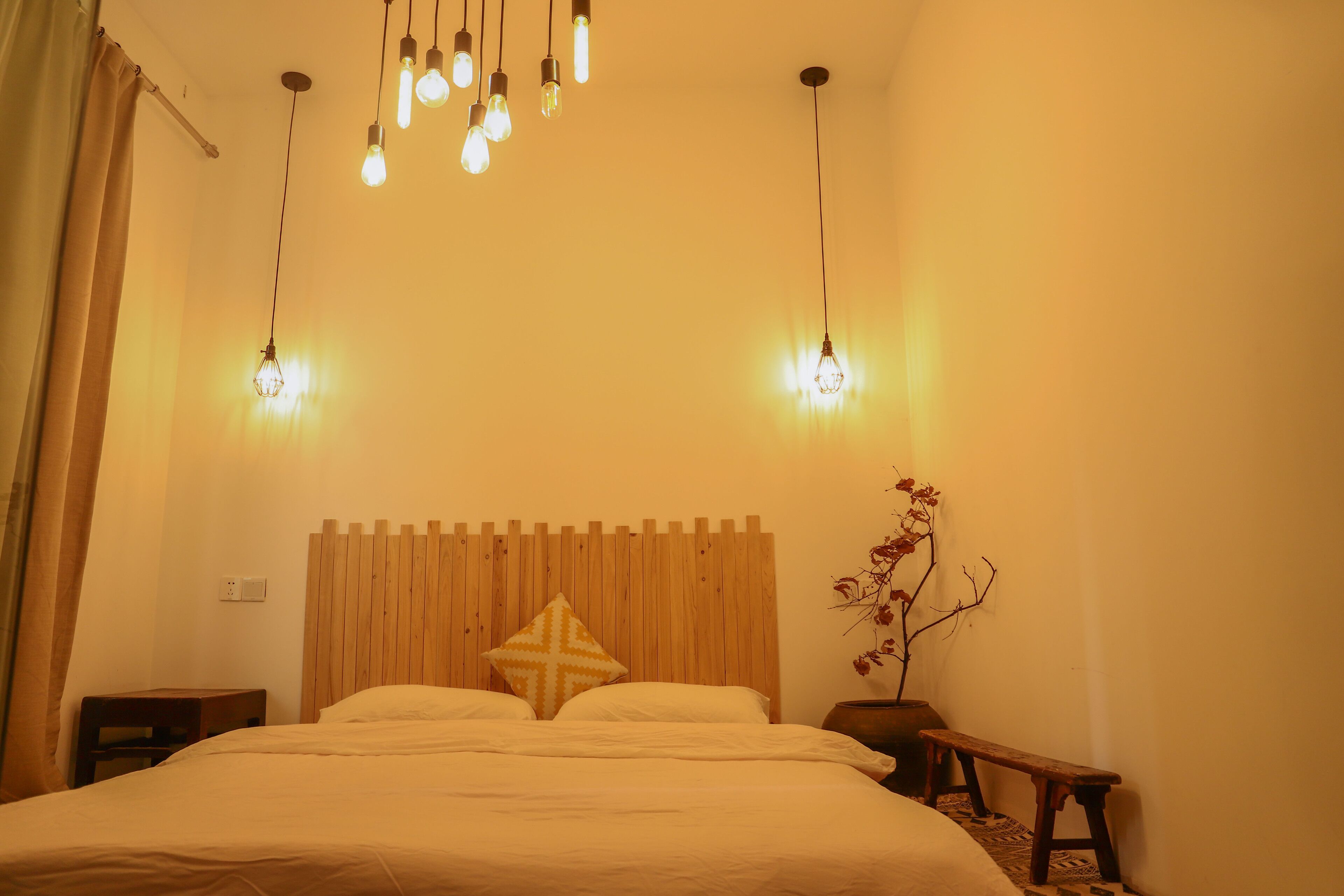 Un dormitorio con cabecera de madera, cama con ropa de cama blanca, mesita de noche, un banco y lámparas colgantes.