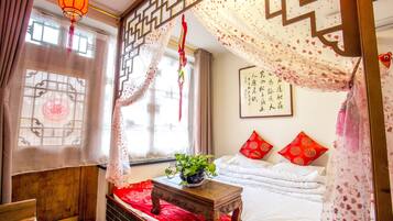 Una habitación tradicional con una cama, una mesita con una planta, un adorno en la pared y una ventana con cortinas.