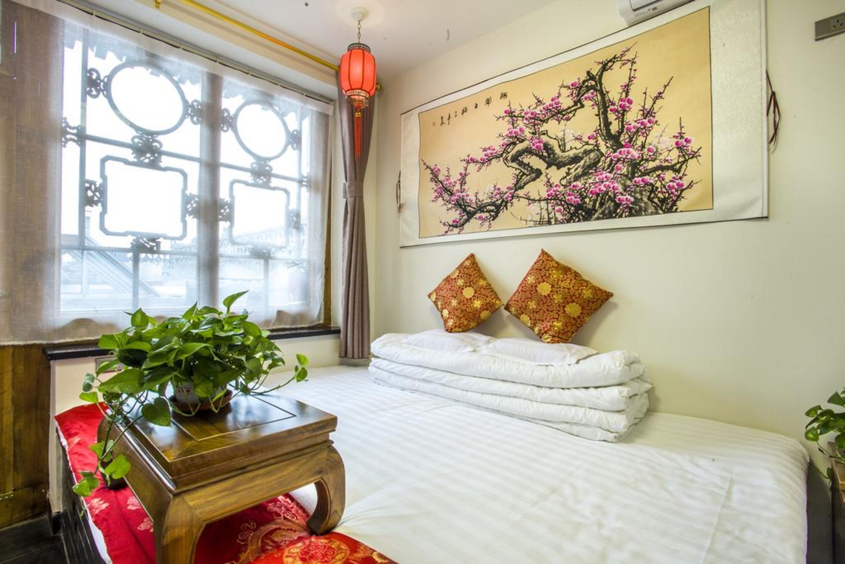 A neatly arranged bedroom with a bed, pillows, a bedside table, a window with curtains, and a wall-mounted painting.