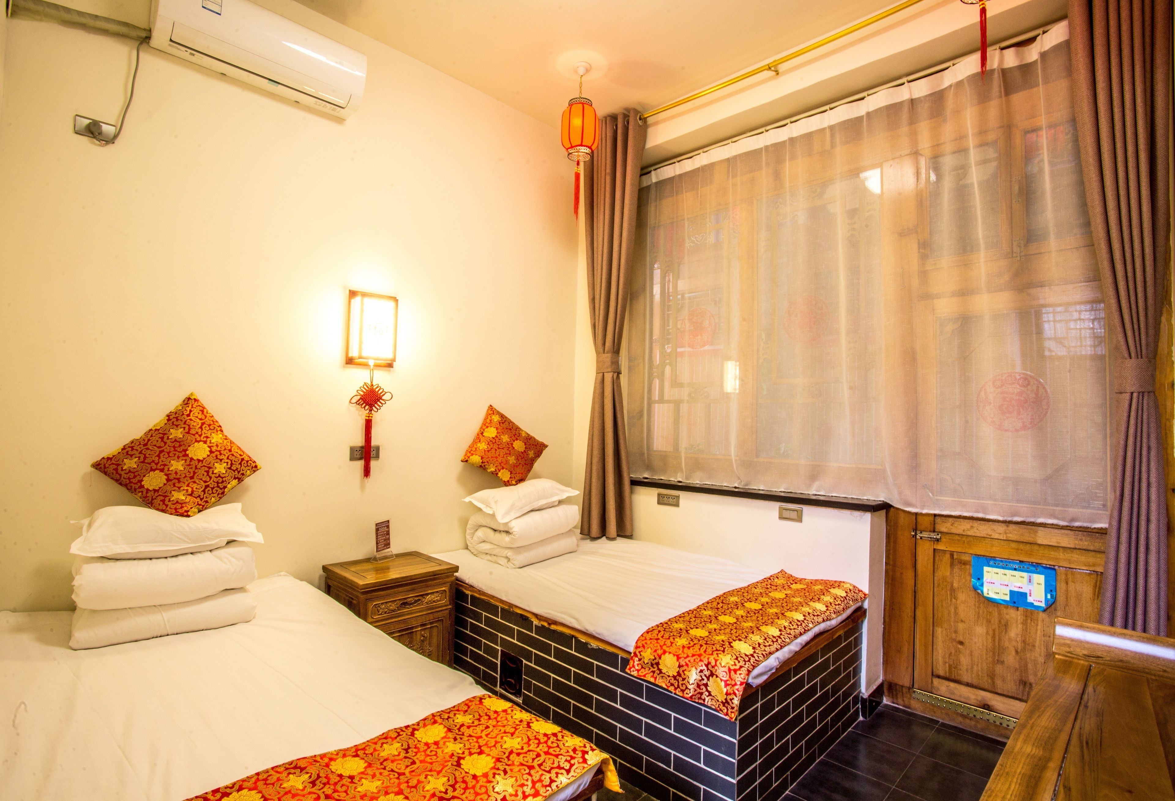 A hotel room with two beds, a window with curtains, an air conditioning unit, and a wooden door with a sign.