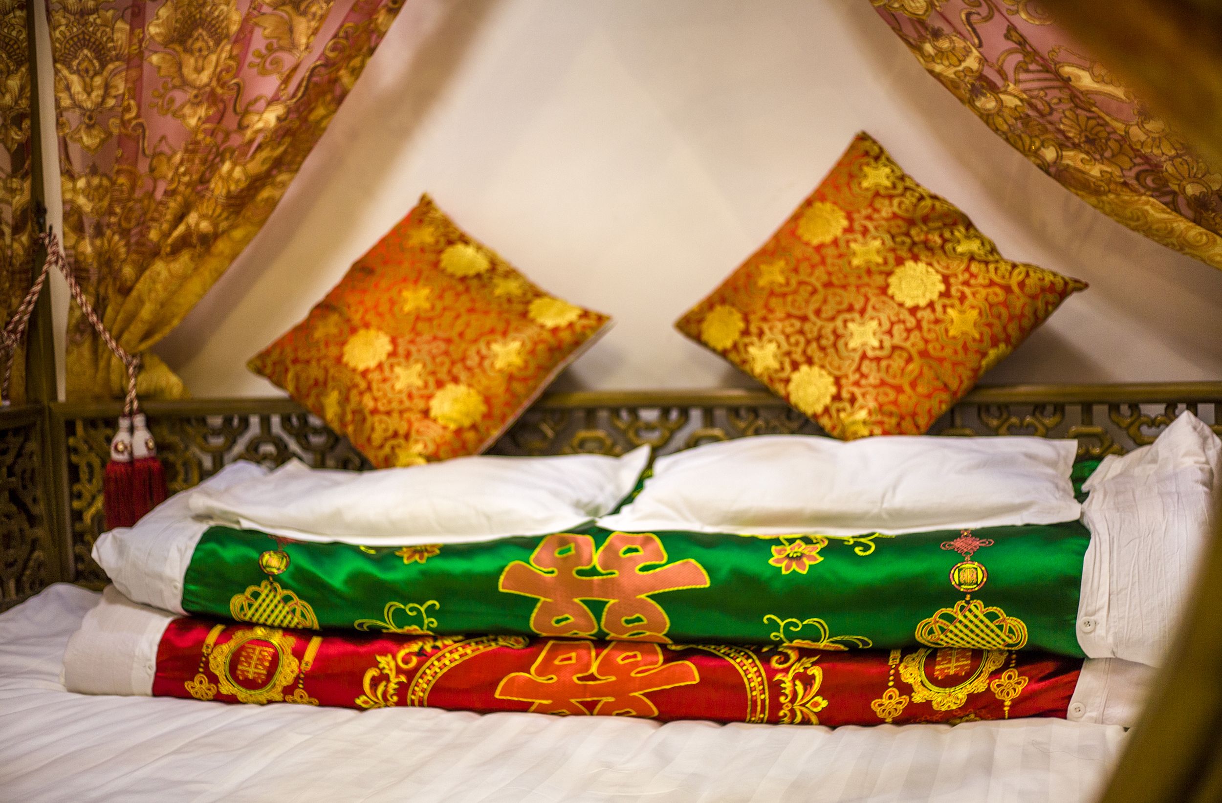 A bed with neatly folded white and green linens, two decorative pillows with golden patterns, and a red and gold embroidered blanket.