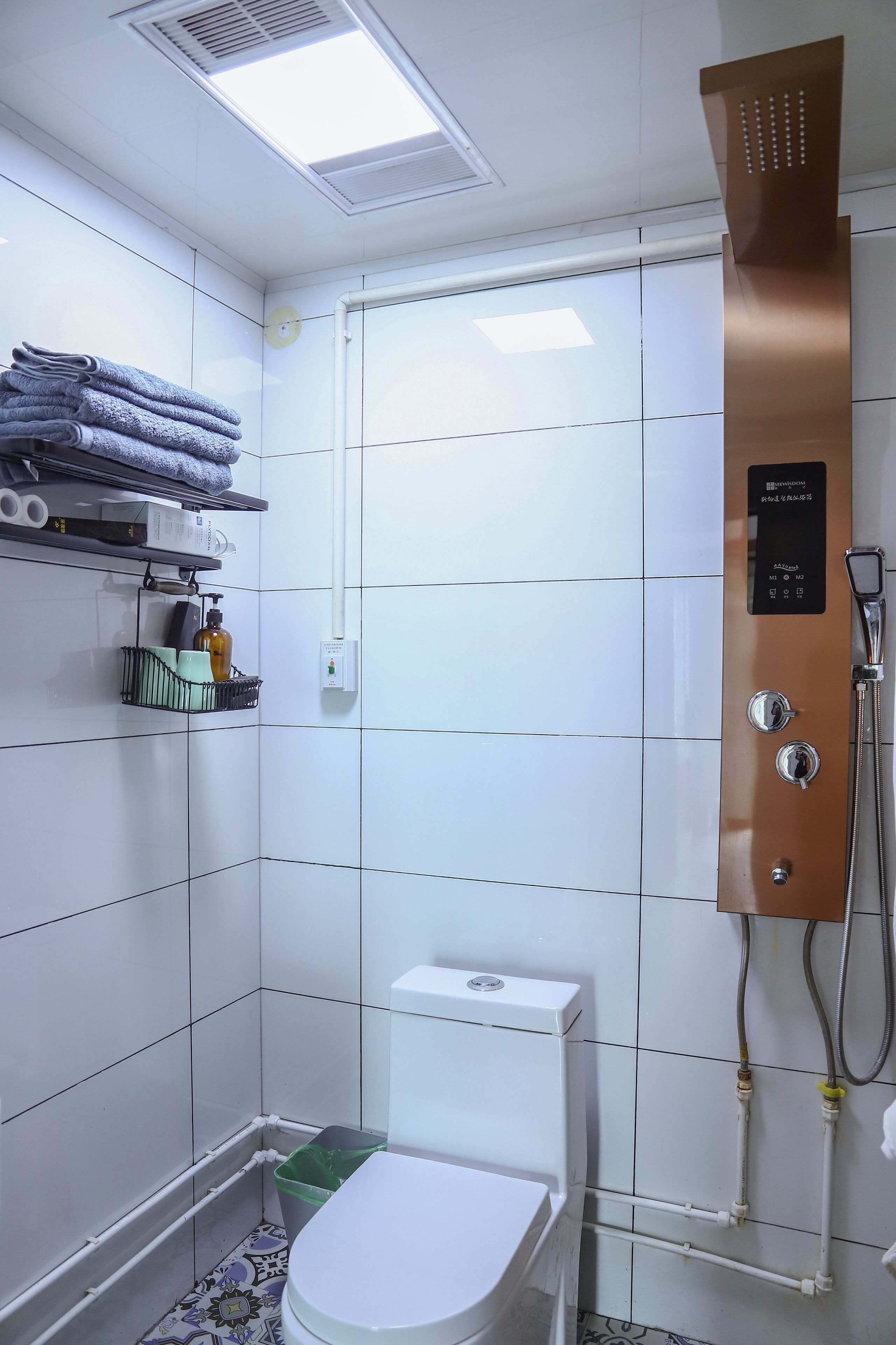 A small, tiled bathroom with a toilet, a shower, and a shelf with towels and toiletries.