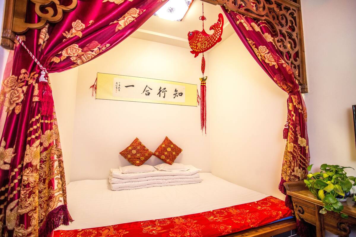 A neatly made bed with white linens and two decorative pillows, a red and gold patterned bedspread, and a wall with a framed sign.