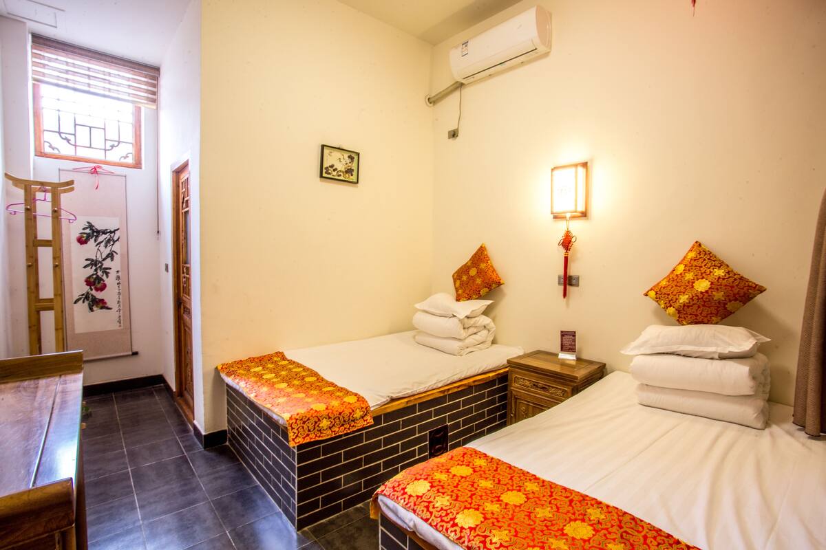 A hotel room with two beds, a wooden desk, a small table, a window with blinds, an air conditioning unit, and a wall-mounted light fixture.