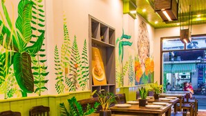 A restaurant with wooden tables and chairs, green walls with plant murals, and a view of the street outside. - Amona Hotel - Hostel (Hue)