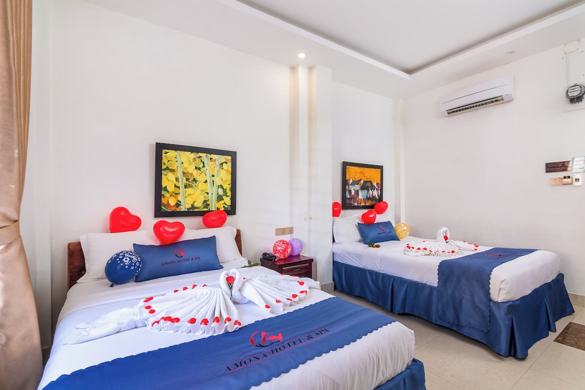 A hotel room with two beds, each with a blue bedspread and white linens, and red heart-shaped pillows. There are wall-mounted air conditioning units and a framed picture on the wall.