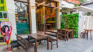 An outdoor seating area with wooden tables and benches, a glass door entrance, and a green wall with plants. - Amona Hotel - Hostel (Hue)