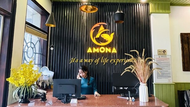 A reception desk with a logo, a sign prohibiting smoking, and a receptionist. - Amona Hotel - Hostel (Hue)
