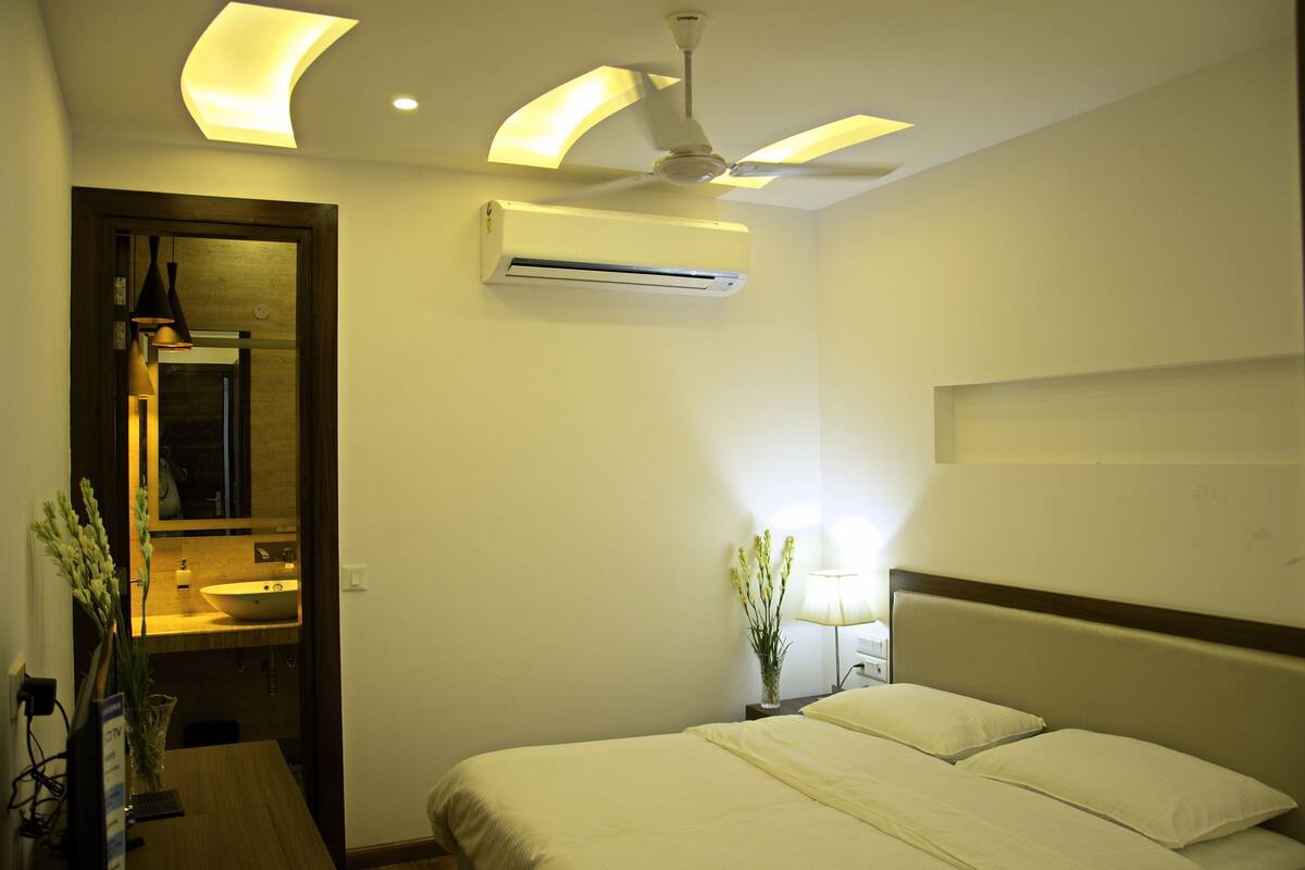 Habitación de hotel con cama, mesita de noche, lámpara, equipo de aire acondicionado, ventilador de techo y baño visible a través de una puerta abierta.