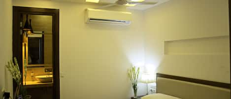 A hotel room with a bed, bedside lamp, air conditioning unit, ceiling fan, and a mirror in the corner.