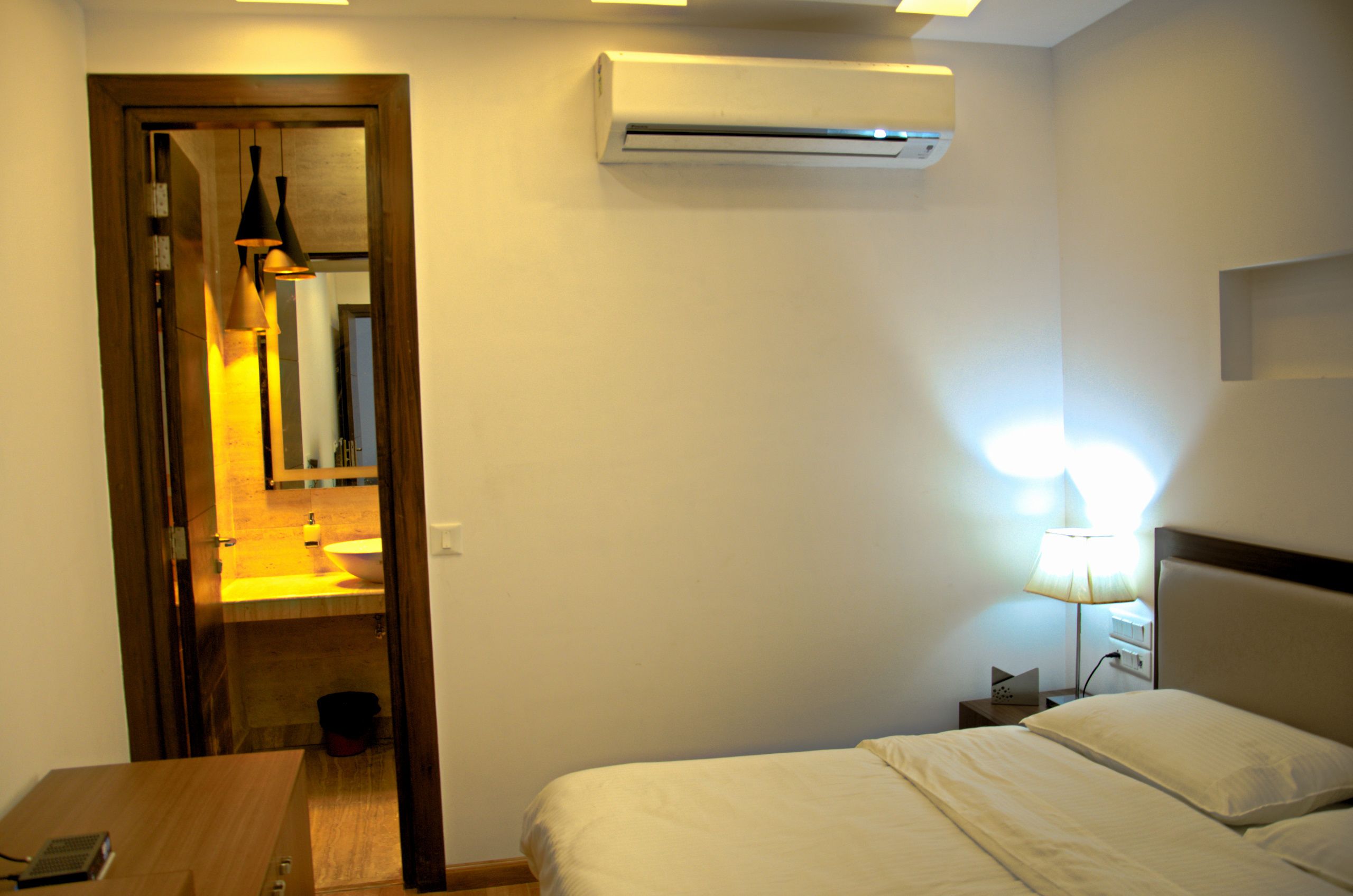 A hotel room with a bed, bedside table, lamp, air conditioning unit, and a bathroom visible through an open door.