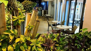 An outdoor seating area with tables and chairs, surrounded by potted plants and a building with a closed shutter. - Grand Hotel Central (Conakry)