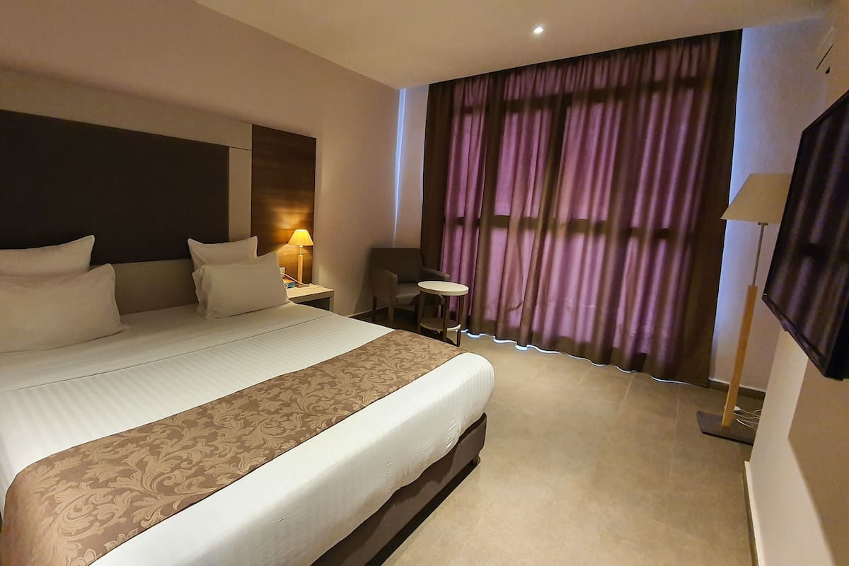 A hotel room with a large bed, a bedside table with a lamp, a chair, and a view of the outside through curtains.
