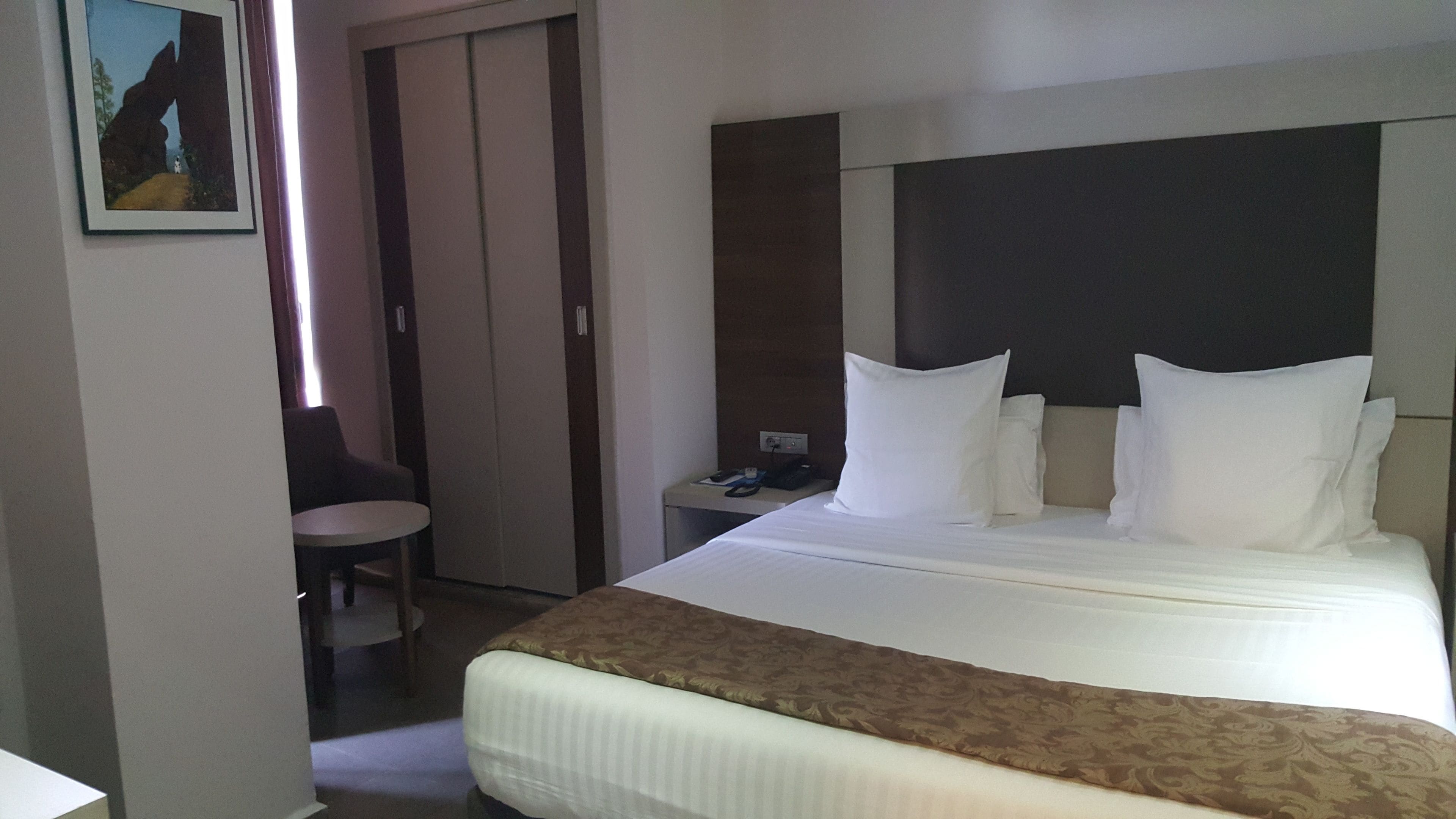 A hotel room with a large bed, a bedside table with a phone and scissors, a chair, and a painting on the wall.