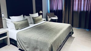 A modern hotel room with a large bed, a chair, a small table, and a window with curtains. - Grand Hotel Central (Conakry)