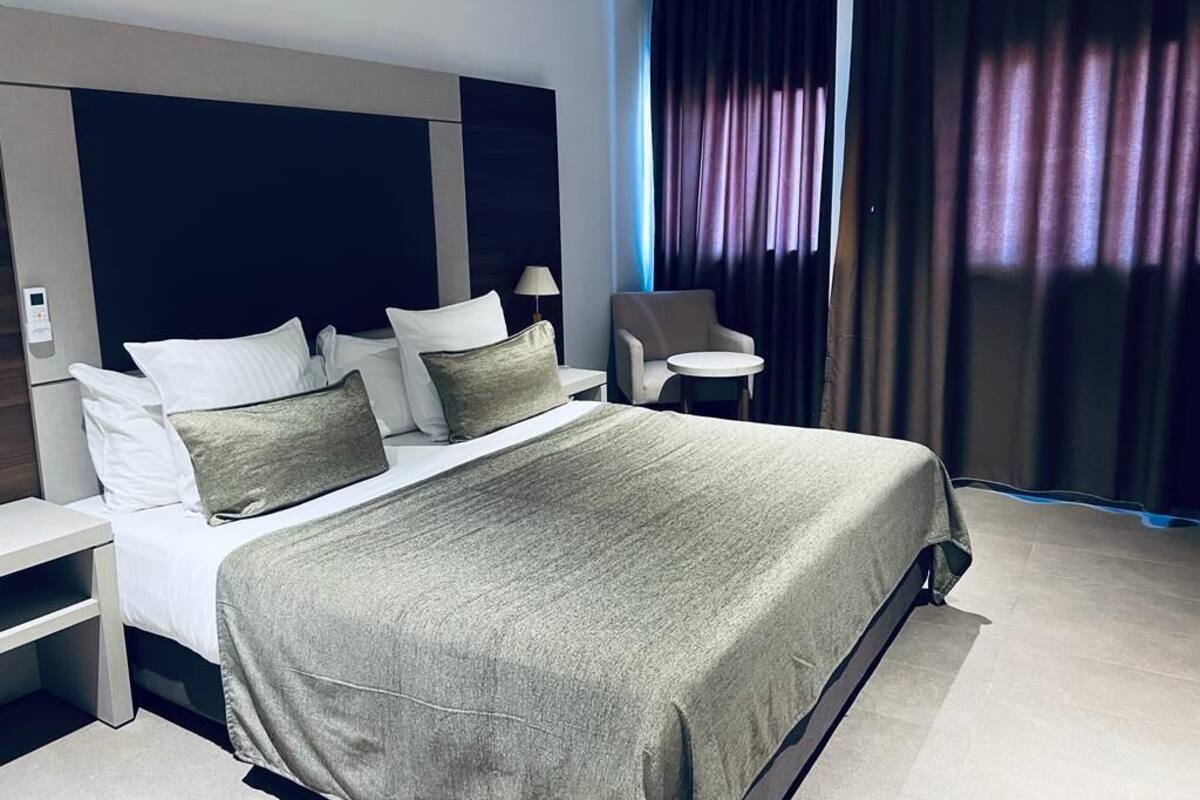 A modern hotel room with a large bed, a chair, a small table, and a window with curtains.