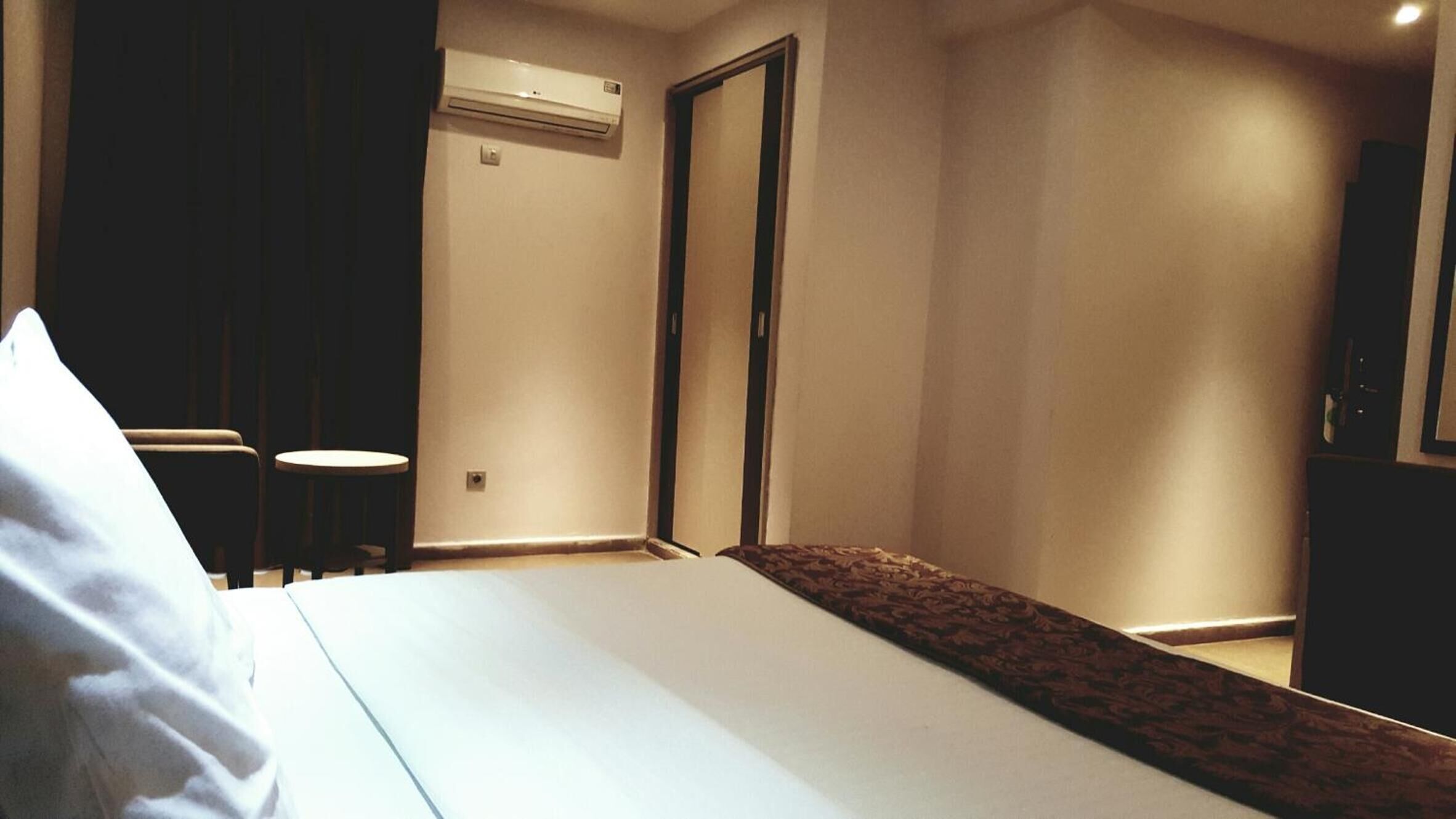 A hotel room with a bed, a small round table, a chair, an air conditioning unit, and a wardrobe.