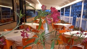 A restaurant with tables and chairs arranged in a row, large windows, and potted plants with flowers. - Hotel Adler (Rimini)