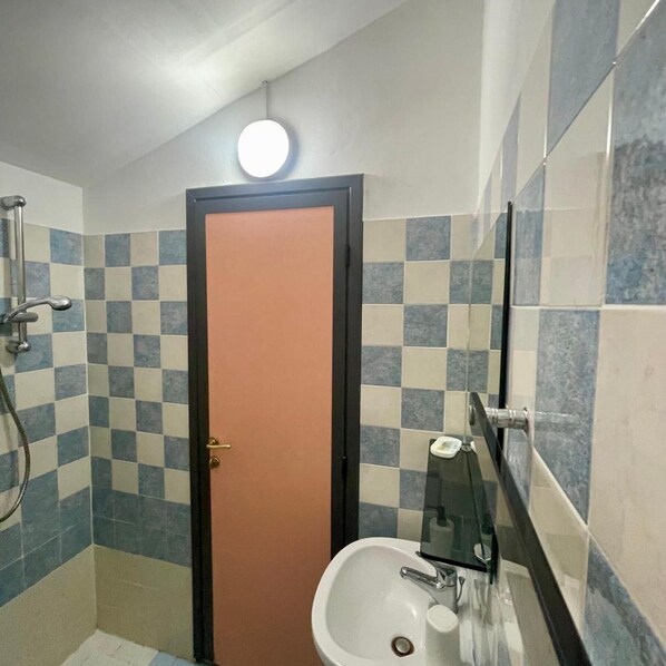 A bathroom with a checkered tile wall, a sink with a mirror, and a shower area. - Hotel Adler (Rimini)