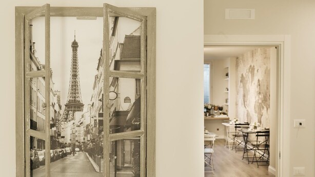 Una habitación con un ventanal que muestra vista a la calle, una zona de comedor con mesas y sillas, y una pared con un mural decorativo.