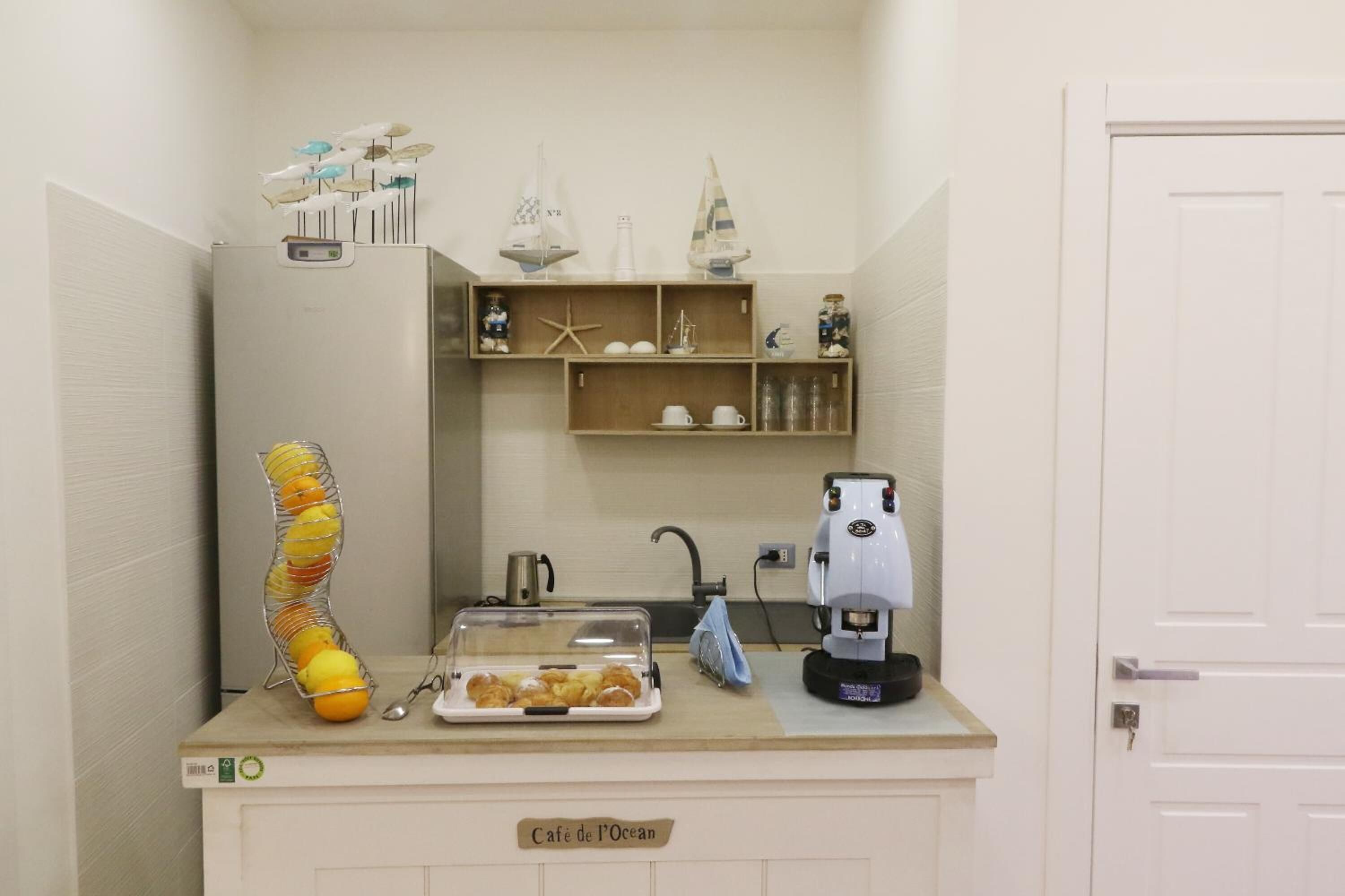 Une petite cuisine équipée d’une machine à café, d’un panier de fruits et d’une pancarte indiquant « Café de l’Océan ».