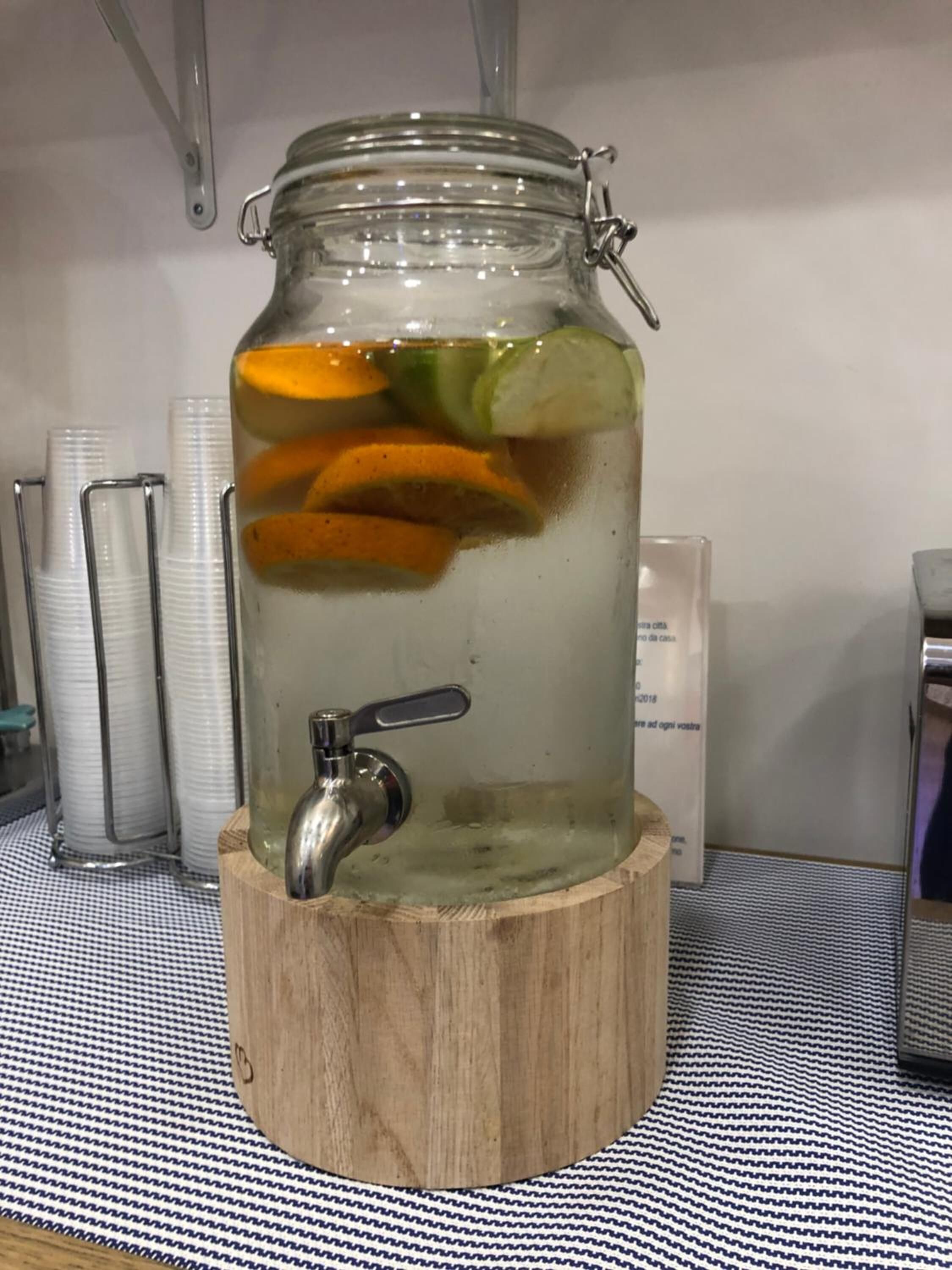 A glass jar with a metal tap filled with water and citrus slices, placed on a wooden stand.