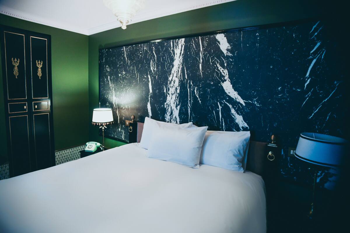 A hotel room with a large bed, a nightstand with a lamp, a telephone, and a wall with a marble-like pattern.