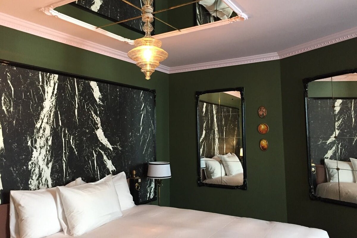 Habitación de hotel con una cama grande, dos espejos en la pared, una araña y un diseño de pared que imita el mármol.