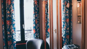Habitación de hotel con un ventanal grande, cortinas florales, una silla, una mesita y un espejo.