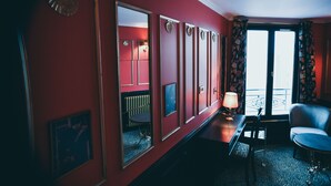 Habitación de hotel con pared roja, un espejo grande, escritorio con lámpara, una silla y vistas al exterior por una ventana.