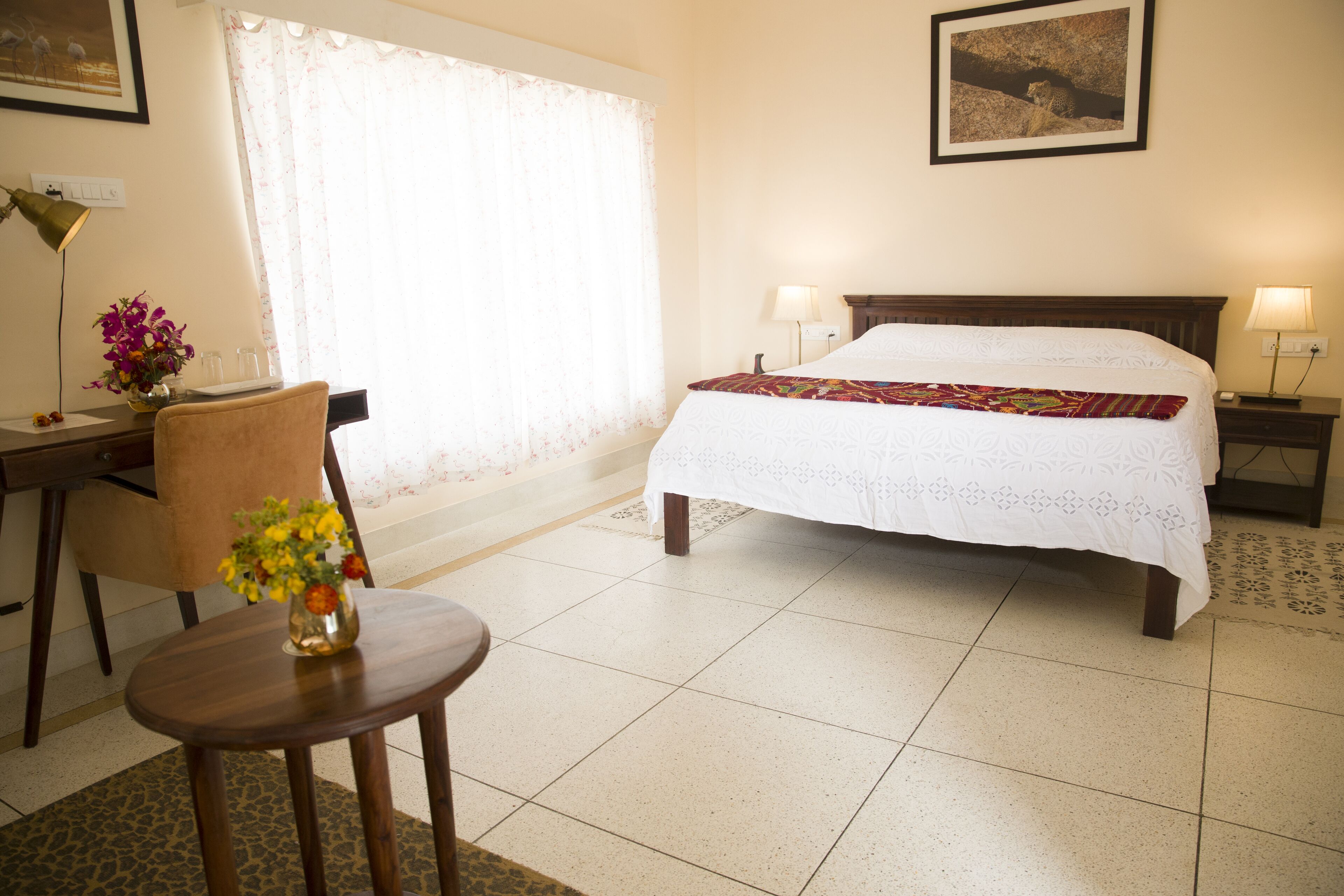 Una habitación de hotel con cama, escritorio con lámpara, silla y un florero con flores.