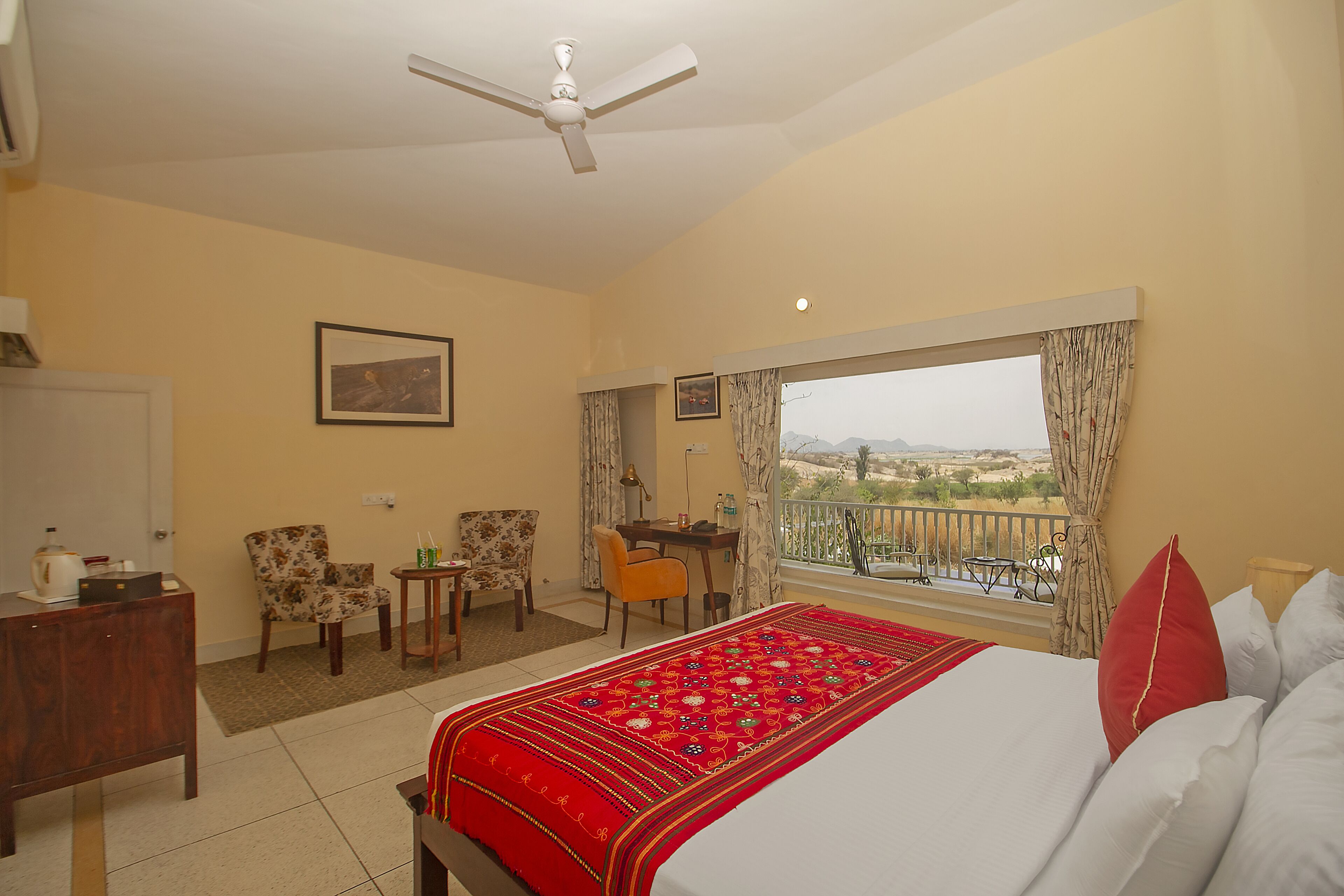 Habitación de hotel con cama, escritorio, dos sillas, una mesita, ventilador de techo y balcón con vistas.