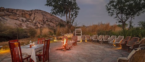 An outdoor dining area with stone flooring, wooden chairs, and a fire pit.