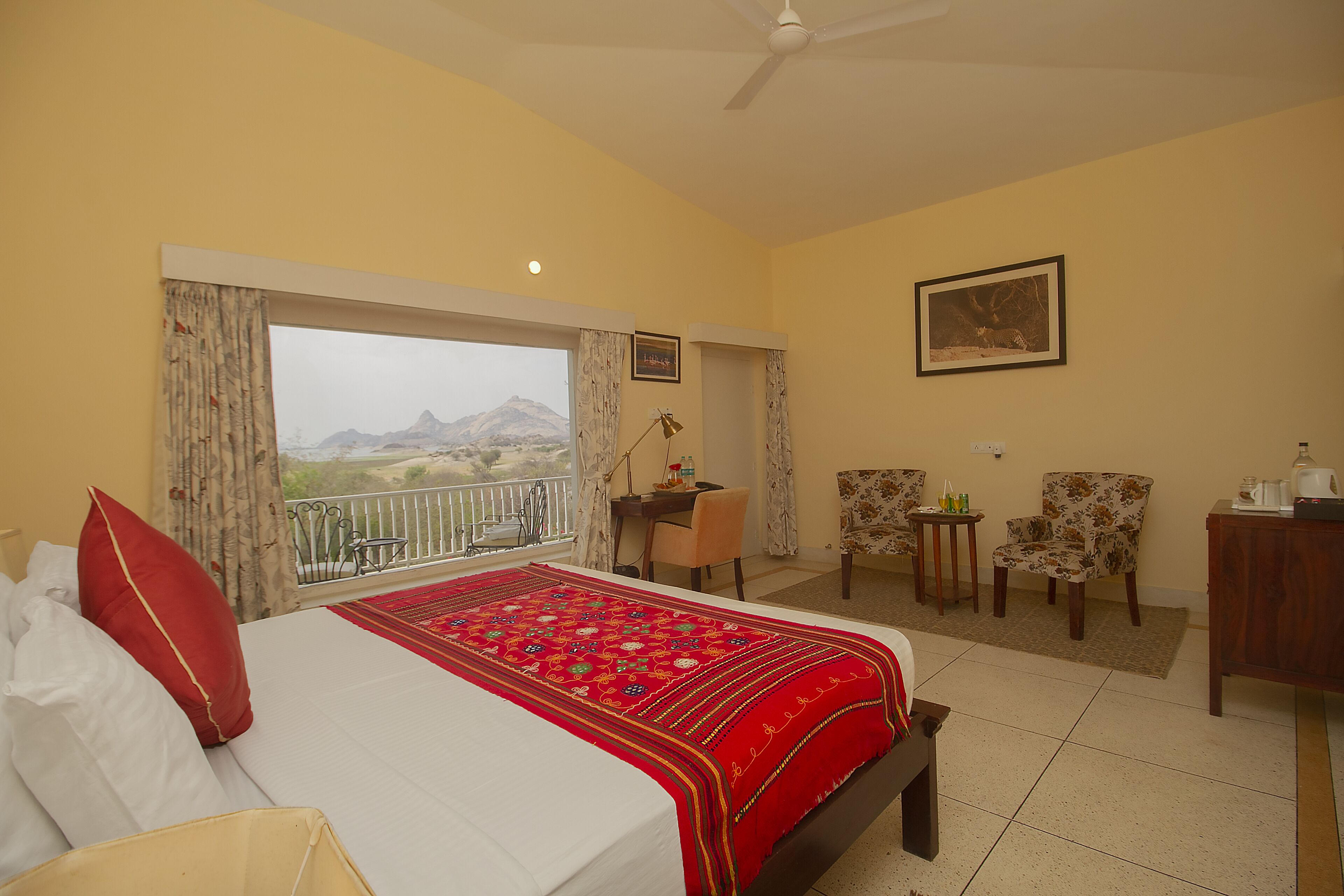 A hotel room with a bed, a desk, two chairs, a balcony with a mountain view, and a painting on the wall.