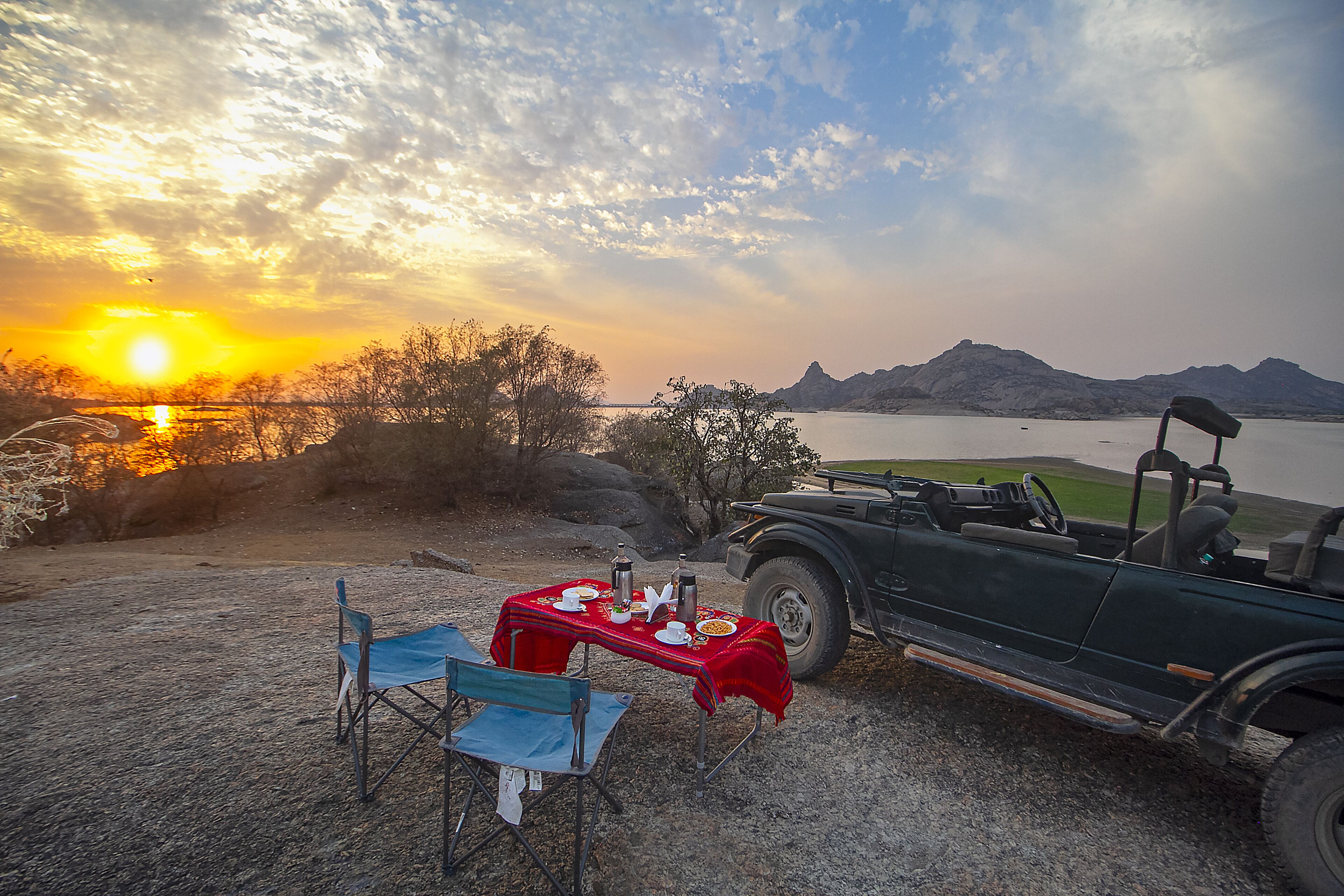 A table set with a meal and a car parked nearby, overlooking a body of water and mountains.