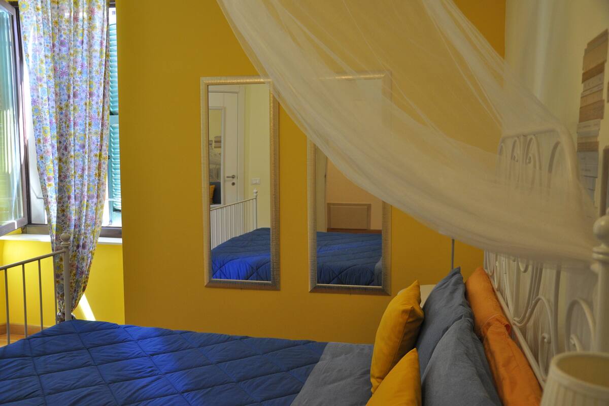 Un dormitorio con cama, paredes amarillas, ventana con cortinas florales y espejo.