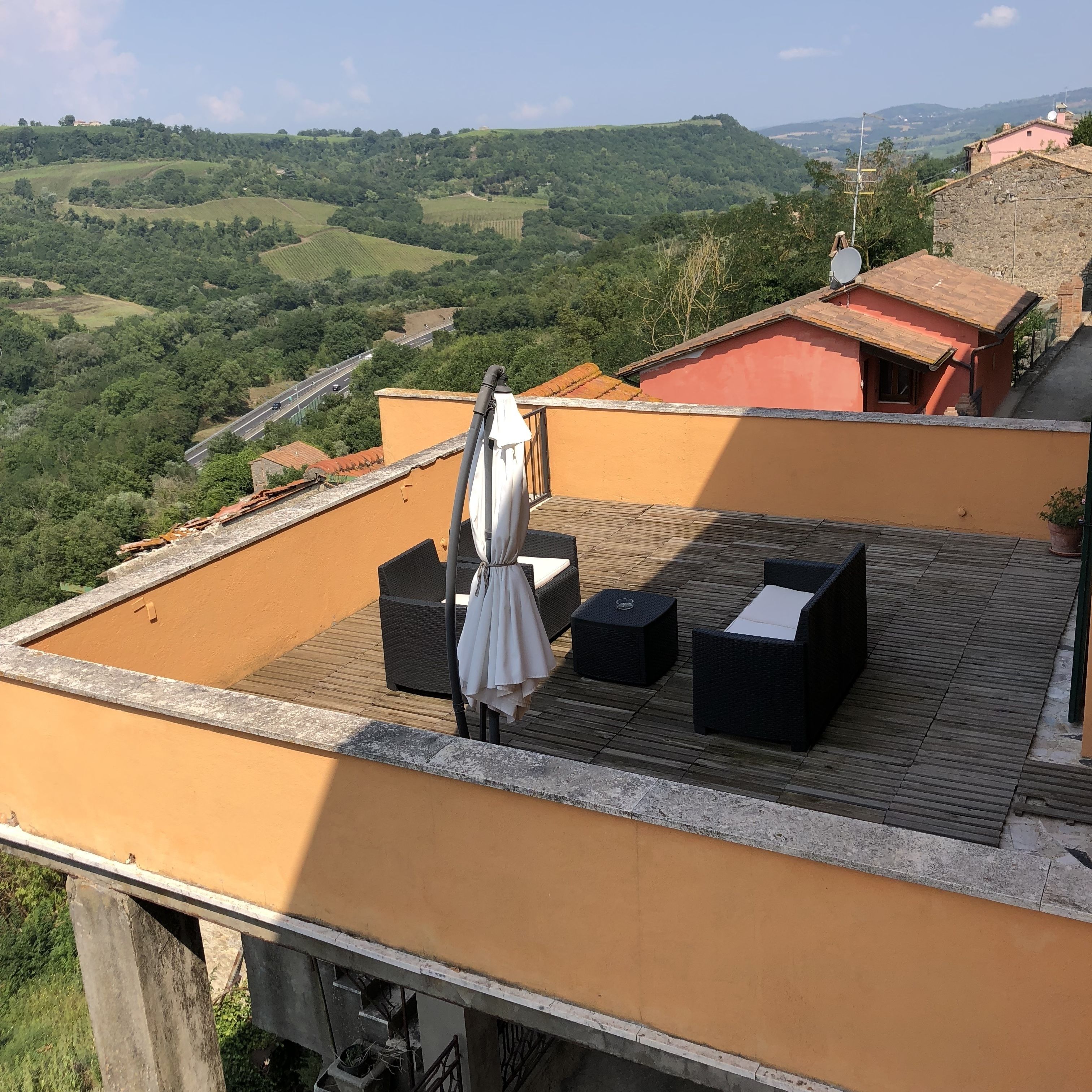 Una terraza en la azotea con mobiliario exterior, un paraguas blanco y una vista panorámica de colinas ondulantes y un edificio lejano.