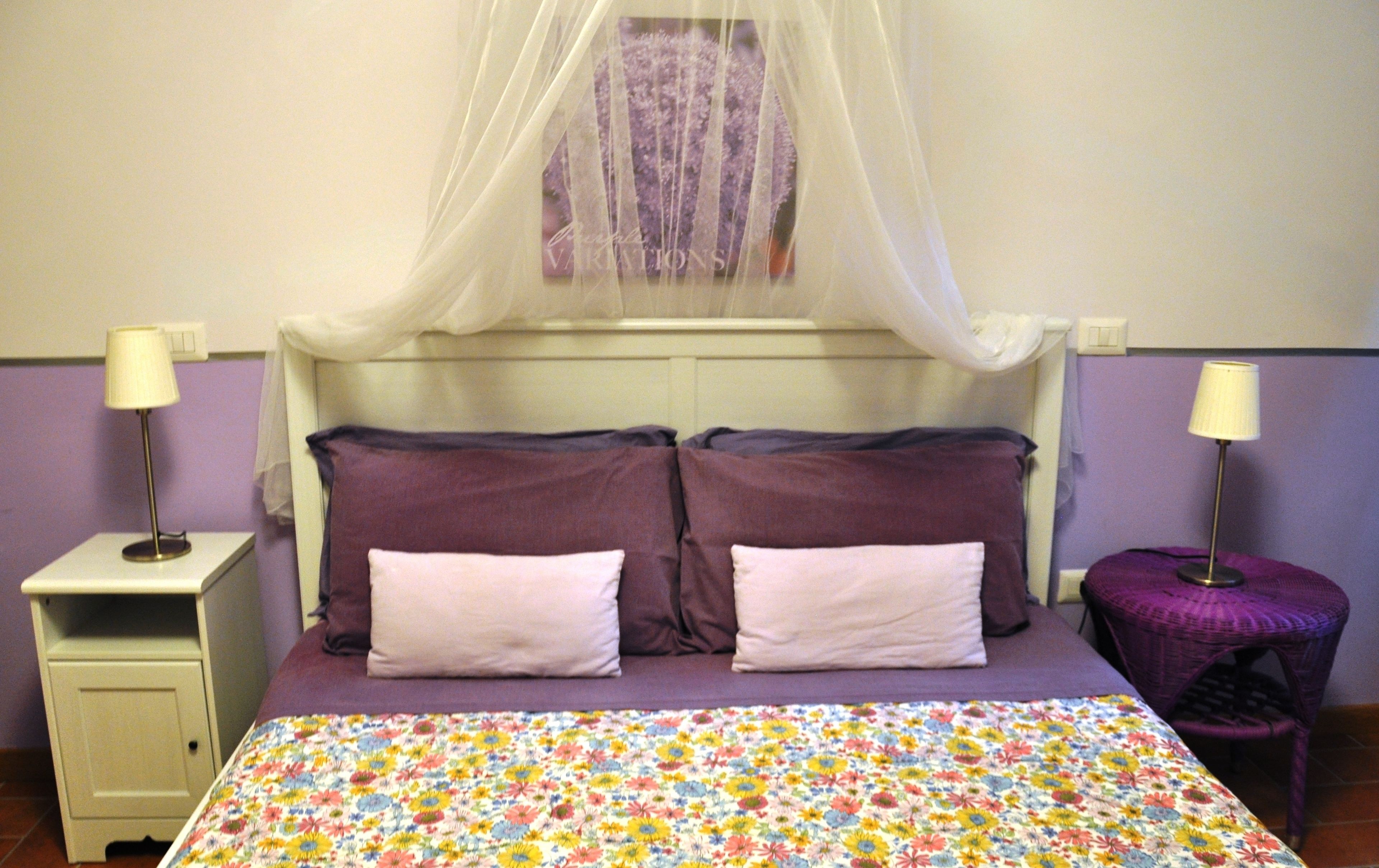A bedroom with a bed, two bedside tables, a nightstand, a purple chair, and a canopy bedspread.
