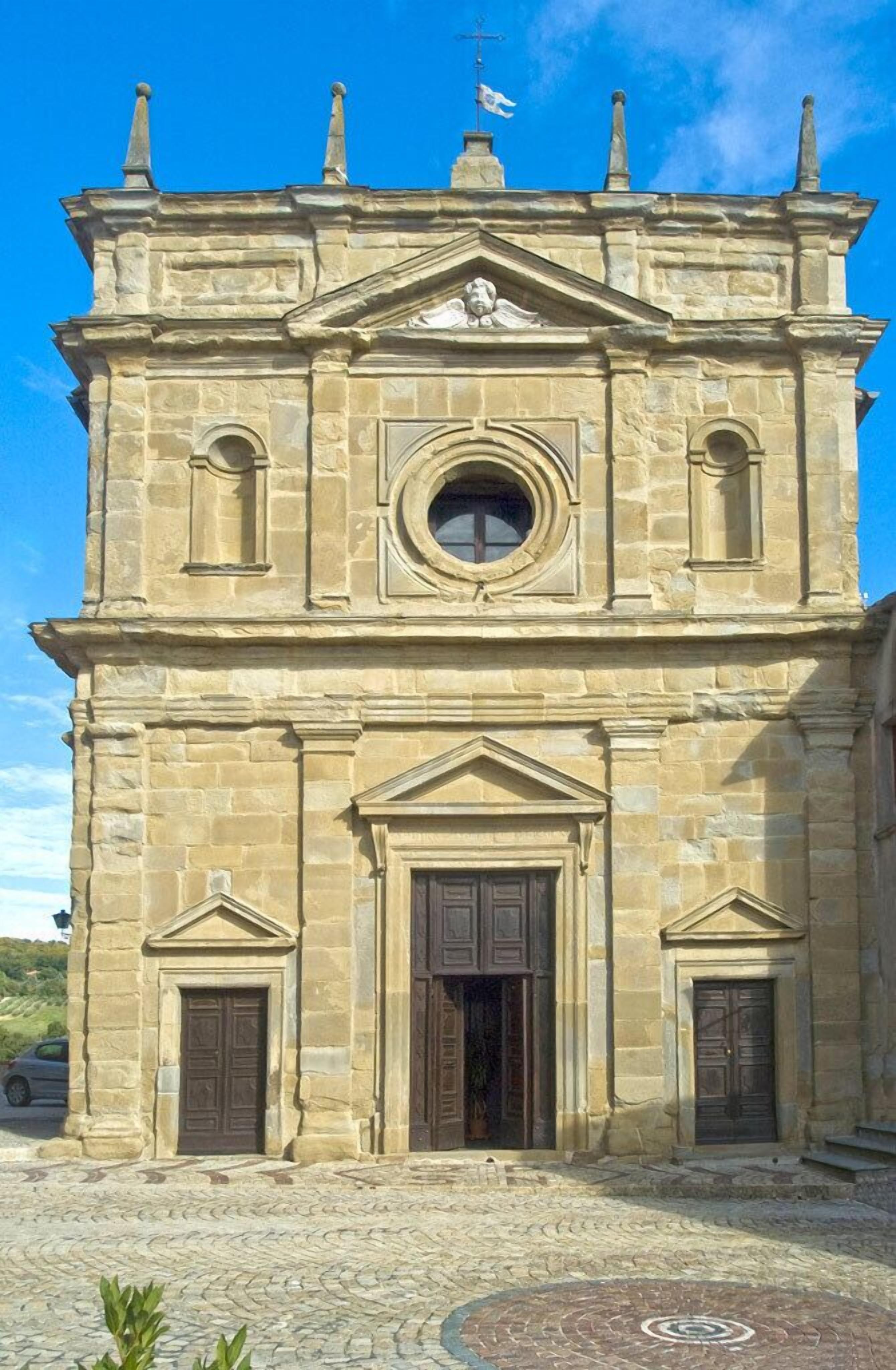 Un bâtiment historique en pierre, doté d’une fenêtre circulaire centrale, flanqué de portes symétriques et surmonté d’une croix.