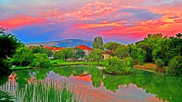 Escena tranquila junto al lago, con el reflejo de árboles y edificios, rodeada de exuberante vegetación y un cielo colorido.