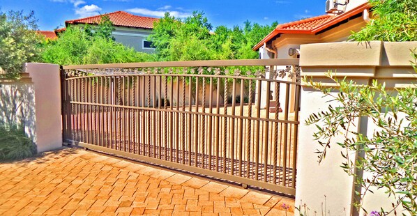 A gated entrance with a metal fence and a brick pathway leading to a building with a red-tiled roof.