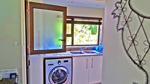 A compact kitchen with a washing machine, a window with a view of greenery, and a decorative wall piece. - Jeanette's Villa at Westlake (Hartbeespoort)
