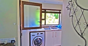 A compact kitchen with a washing machine, a window with a view of greenery, and a decorative wall piece. - Jeanette's Villa at Westlake (Hartbeespoort)