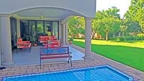 A covered outdoor area with a pool, seating, and a view of a well-maintained lawn and trees. - Jeanette's Villa at Westlake (Hartbeespoort)