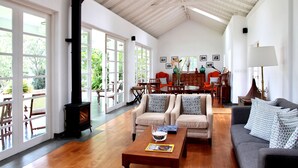 A spacious living room with a high ceiling, large windows, a fireplace, and a dining area with wooden furniture. - The Planters House (Koslanda)