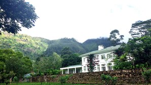 A large white building with a stone wall, surrounded by lush greenery and hills. - The Planters House (Koslanda)