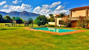 A swimming pool surrounded by a well-maintained lawn, a playground, and a building with a red roof. - Key West Condo Apartment (Hartbeespoort)