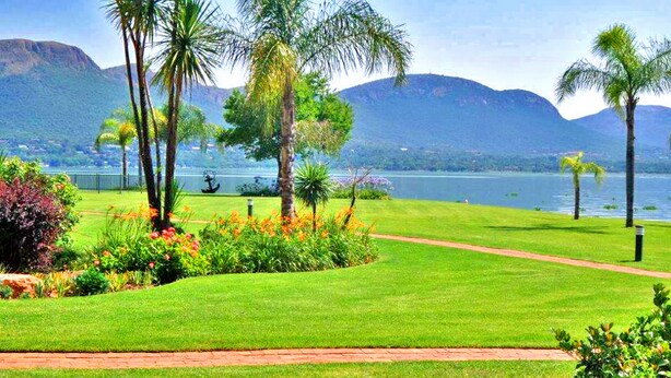 A well-maintained grassy area with palm trees and a path leading to a body of water, with mountains in the background. - Key West Condo Apartment (Hartbeespoort)