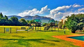 A playground with slides and climbing structures, a fenced area, and a swimming pool. - Key West Condo Apartment (Hartbeespoort)