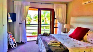 A bedroom with a bed, a TV, a window with curtains, and a view of greenery outside. - Key West Condo Apartment (Hartbeespoort)